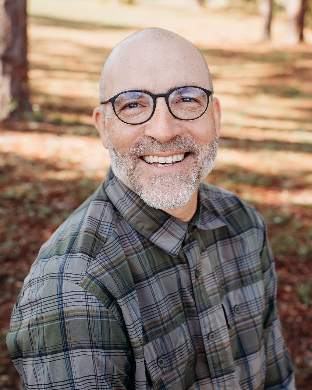 Portrait of a smiling man with glasses and a gray beard in a plaid shirt outdoors
