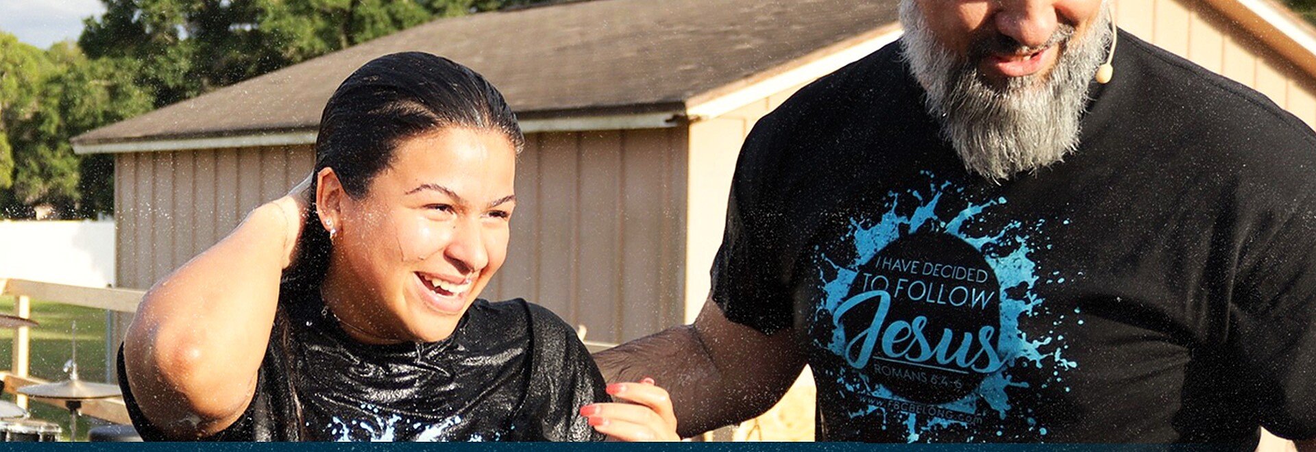 A smiling woman being baptized, standing beside a bearded man in a Jesus t-shirt