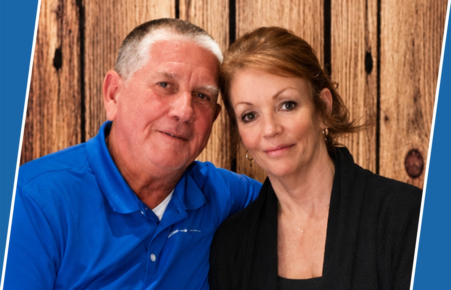 Smiling older man in a blue polo beside a woman with auburn hair against a wooden wall