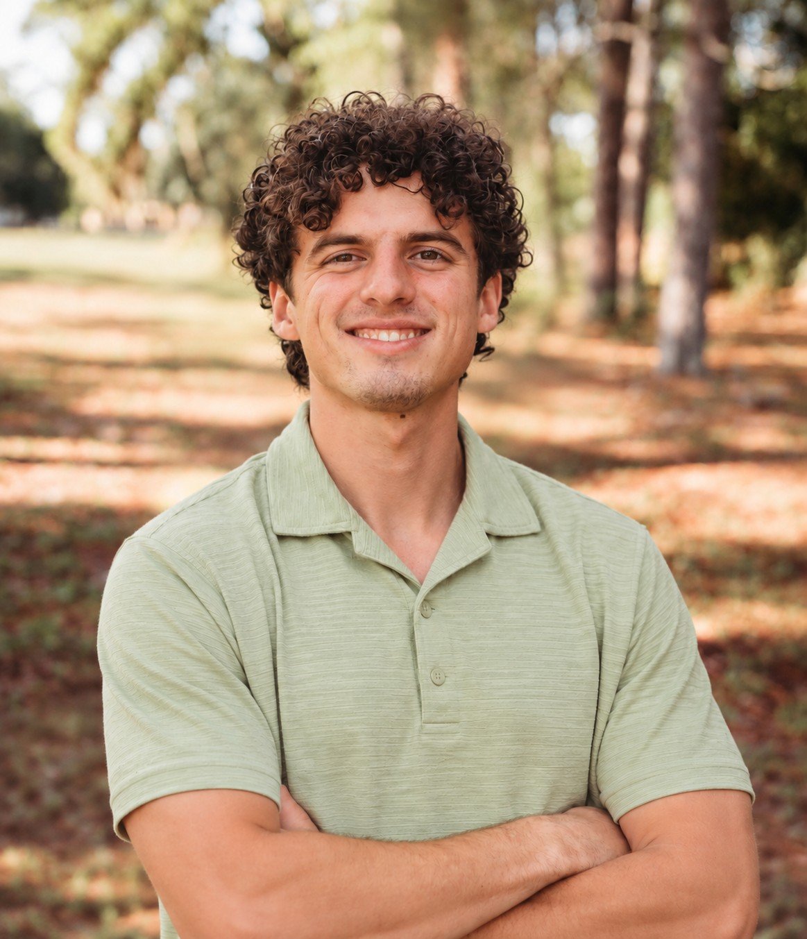 Portrait of a smiling young man with curly hair and arms crossed, wearing a green polo outdoors