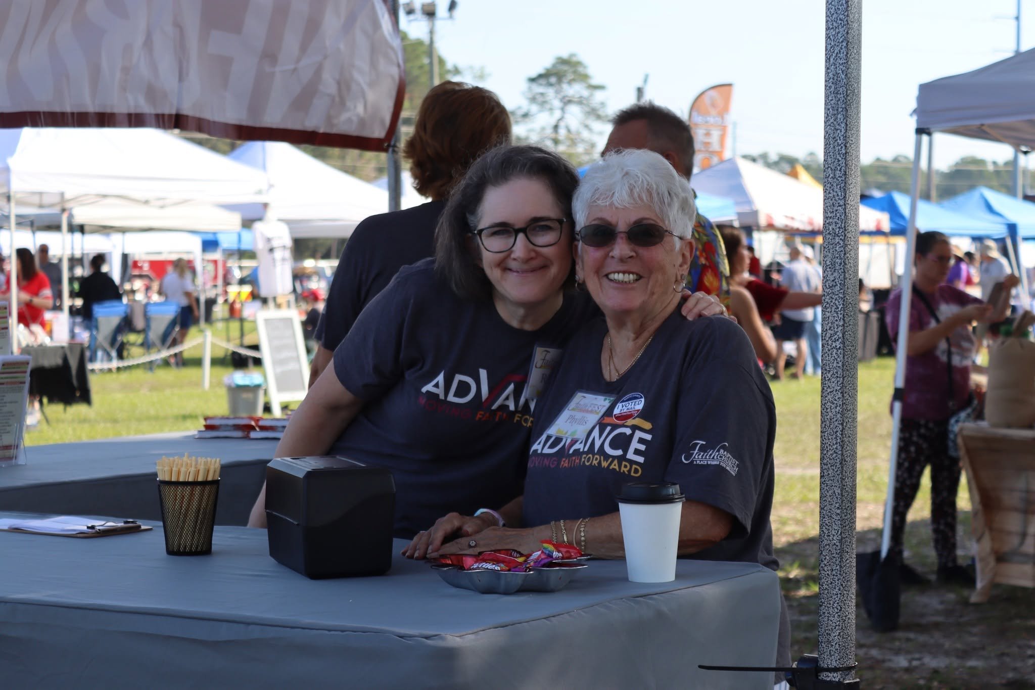 Two smiling women at a table at an outdoor festival with tents behind them