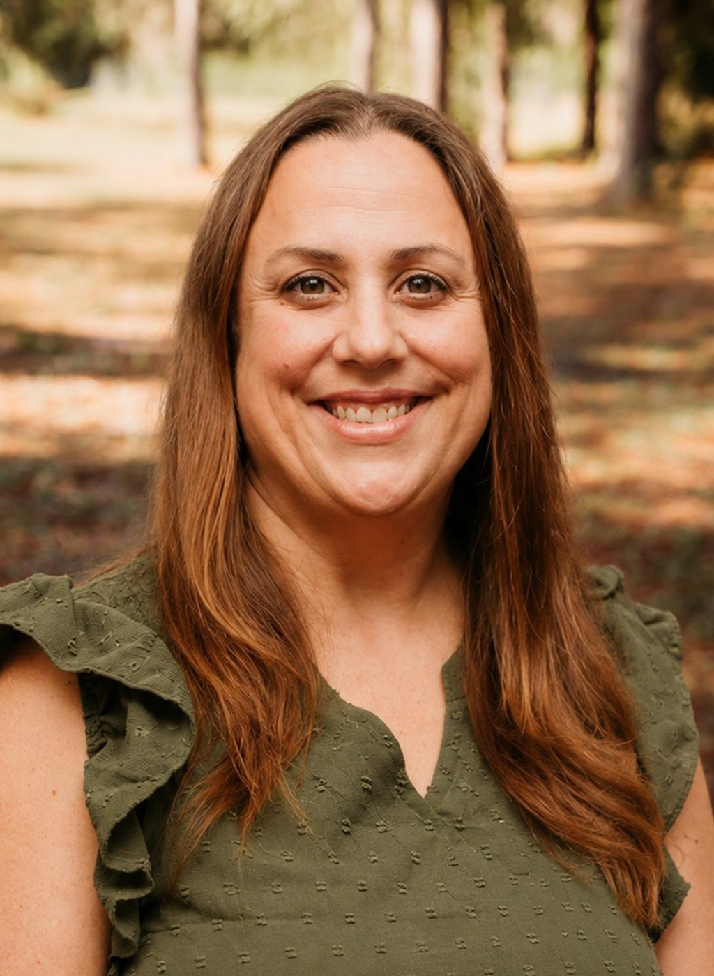 Portrait of a smiling woman with long brown hair in an olive top outdoors
