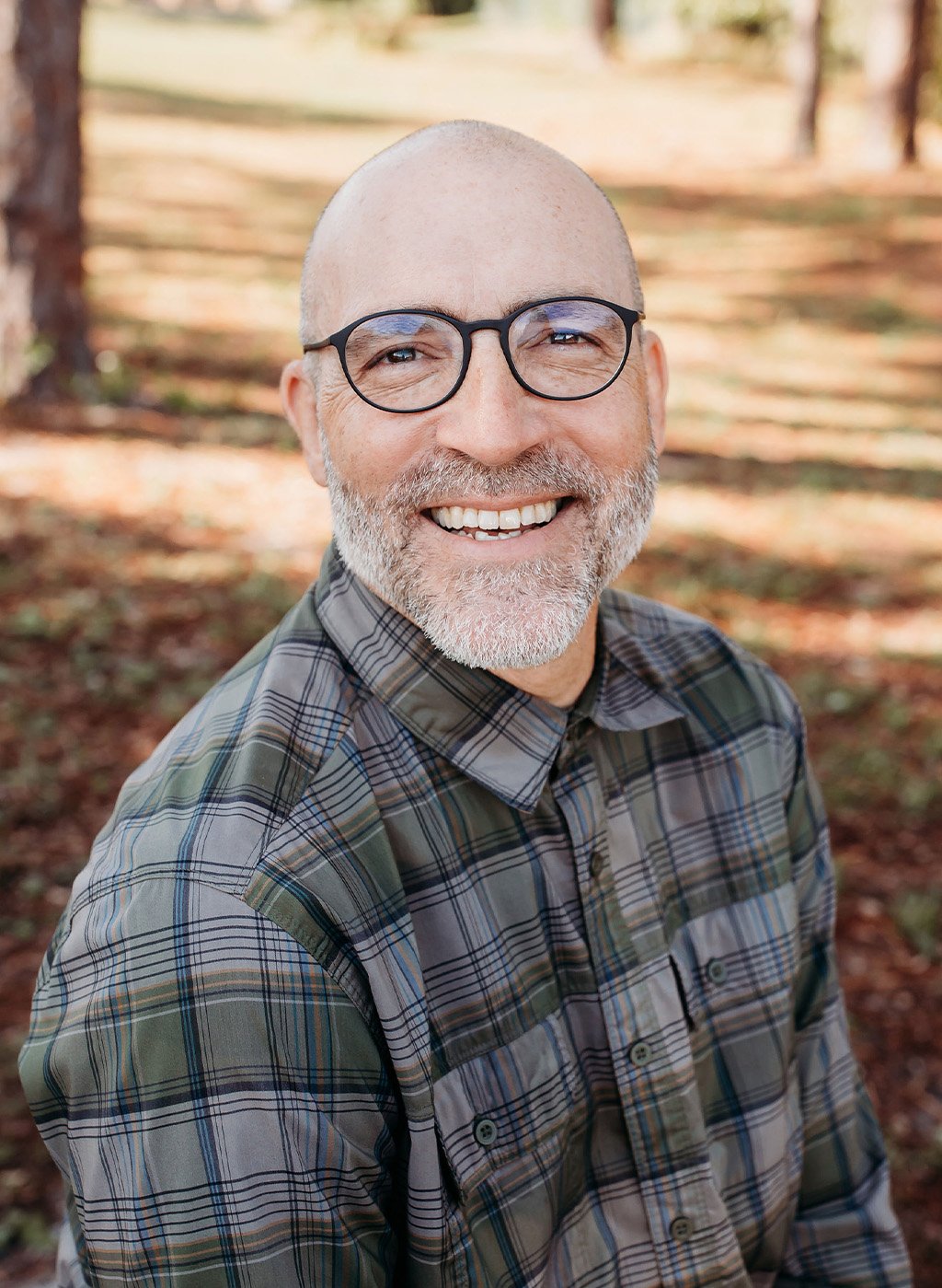 Portrait of a smiling man with glasses and a gray beard in a plaid shirt outdoors